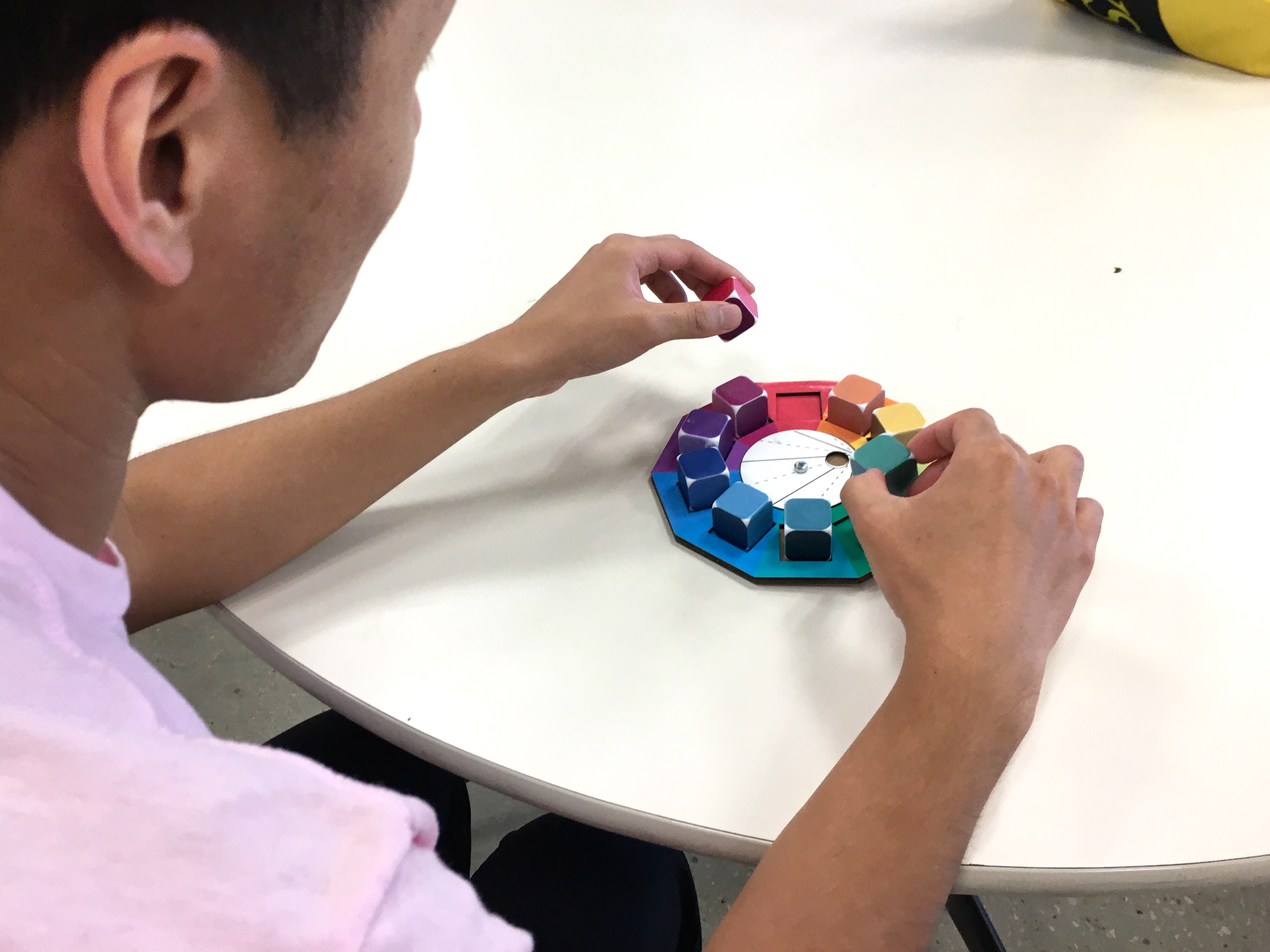 A person picks up colorful dice on a cardboard prototype of a spinning color wheel, testing it.
