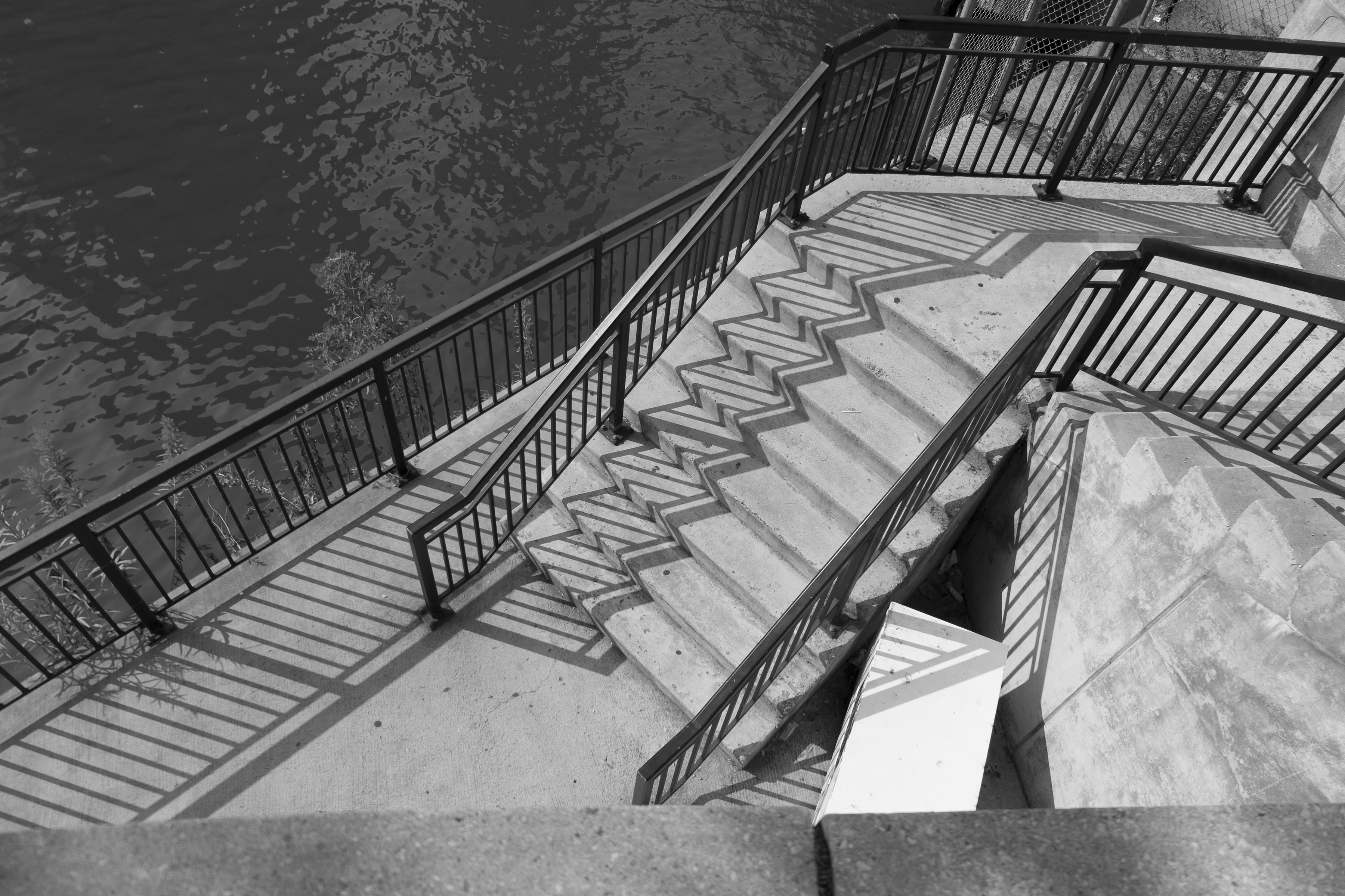 A photograph of a staircase down to a riverside. The stair railings are casting stark shadows on the stairs.
