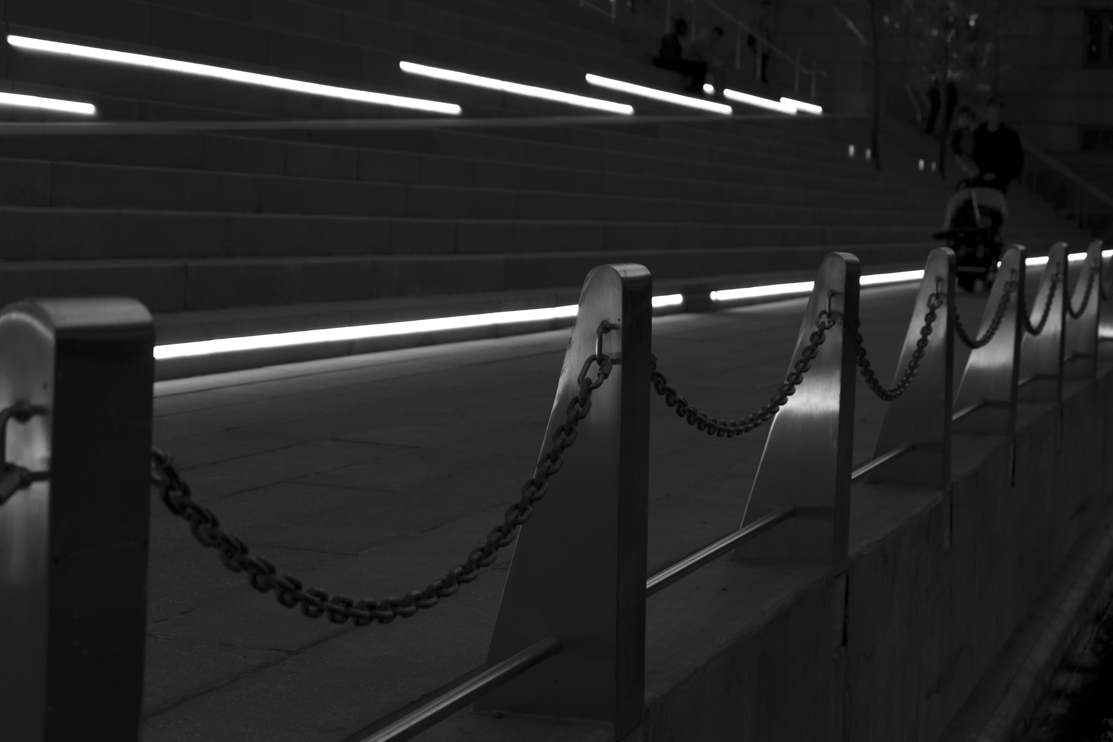 A dark photograph of a chain railing along the riverfront, with bars of bright light on staircases in the background. Perspective disappears into the right background.